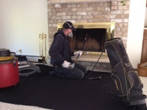 Technician wearing protective gear performing chimney sweeping services with vacuum and rods in front of a fireplace.